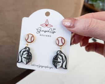 May include: A pair of baseball-themed earrings. Each earring features a baseball stud with a red and white design, connected to a black and white knight helmet charm. The earrings are displayed on a white card with the text "Ginger Up Designs".