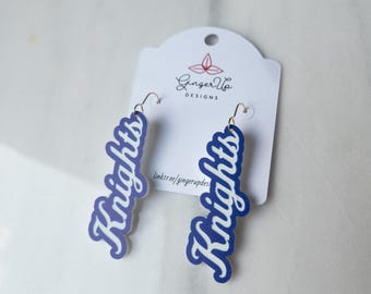 May include: Blue and white 'Knights' earrings with gold-tone hooks. The earrings are in a script font and are presented on a white card with the 'Ginger Up Designs' logo and a website link. The earrings are a stylish accessory.