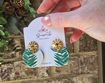 May include: A pair of Ginger Vo Designs earrings. Each earring has a gold-coloured, flower-shaped stud and a white, half-circle pendant with a green fern leaf design. The earrings are displayed on a white card with the Ginger Vo Designs logo.