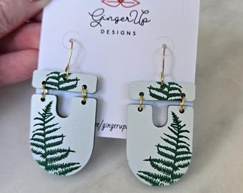 May include: Pale blue earrings featuring a fern leaf design. The earrings have gold-coloured hooks and are attached to a white card with the text "Ginger Up Designs". The fern is painted in green.