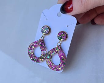 May include: A pair of glittery pink and gold earrings. The earrings have a teardrop shape with a gold-coloured border and a smaller, round stud. The earrings are on a white card.