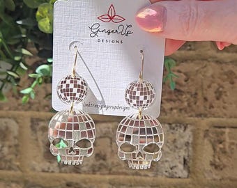 May include: A pair of drop earrings with a disco ball design. The earrings have a skull shape at the bottom and a smaller disco ball above. The earrings are suspended from a gold-coloured hook. The earrings are on a white card with the text "Ginger Up Designs".