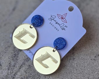 May include: Circular gold-coloured earrings featuring a stylized letter "L" design. Each earring is attached to a small, dark blue, glittery stud. The earrings are displayed on a light purple card with the text "Ginger Up Designs".