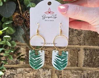 May include: Gold hoop earrings featuring a white and green fern leaf design. The earrings are displayed on a white card with the text "Ginger Up Designs". The earrings are held up by a person's hand.
