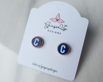 May include: A pair of round stud earrings with a gold-coloured border. Each earring features a blue background with a white letter "C". The earrings are displayed on a white card with the text "Ginger Up Designs" and a link to their website.