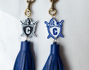 May include: Two navy blue tassel keyrings. Each keyring features a gold-coloured clasp, a shield-shaped emblem with a knight's helmet and crossed swords, and the letter "C". The tassels are made of fringed leather.