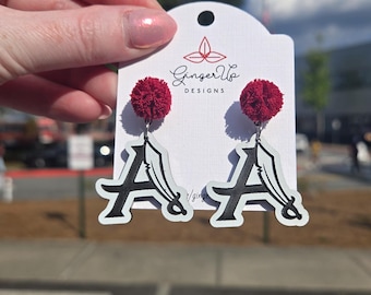 May include: Earrings with a black 'A' design and white outline, each topped with a small, round, burgundy pom-pom. The earrings are presented on a white card with the "Ginger Up Designs" logo.