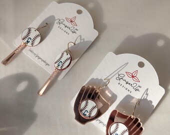 May include: Baseball-themed earrings. One pair features a baseball and a bat, the other a baseball in a mitt. Each earring has a white baseball with blue stitching and the letter "C". The earrings are on white cards with the text "Ginger Up Designs".