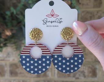 May include: Dangle earrings featuring a gold-toned textured circular stud attached to a larger wooden circular drop. The drop displays a red and white striped pattern on the top half and a blue field with white stars on the bottom half.
