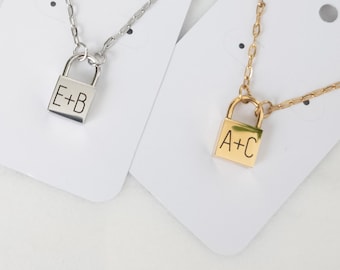 May include: Two necklaces featuring padlock pendants. One is silver, engraved with "E+B", the other gold, engraved with "A+C". Both pendants are square and attached to fine chain necklaces. The pendants are displayed on white cards.