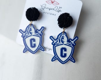 May include: Blue and white shield-shaped earrings featuring a letter "C" and crossed swords, with black pom-poms. The earrings hang from a white card with the text "Ginger Up Designs".