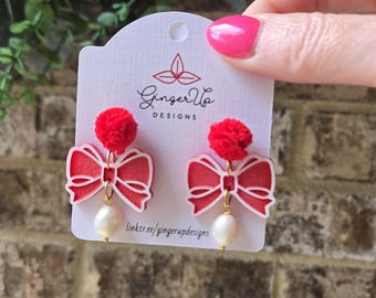 May include: Red bow earrings with a white outline, a red pom-pom, and a dangling pearl. The earrings are displayed on a white card with the text "Ginger Up Designs" and a website address. The background is a brick wall.