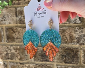 May include: A pair of teal glitter earrings with orange leaf-shaped accents. The earrings are suspended from gold-coloured posts and are displayed on a white tag with the text "Ginger Up Designs".