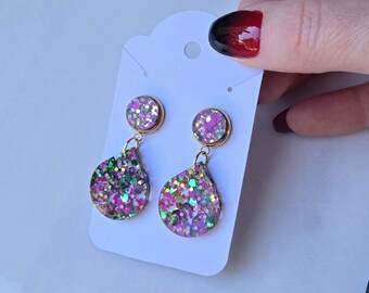 May include: Dangle earrings with a gold-coloured setting. Each earring has a round stud and a teardrop-shaped dangle, both filled with pink, green, and gold glitter. The earrings are presented on a white earring card.