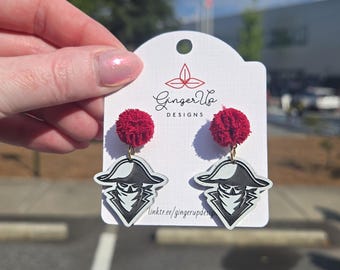 May include: Earrings featuring a red pom-pom and a pirate design. The earrings are displayed on a white card with the text "Ginger Up Designs" and a website link. The earrings are held up by a hand.
