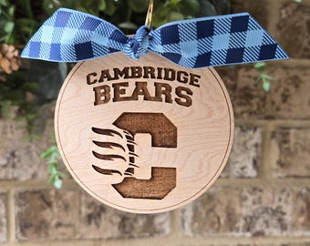 May include: Circular wooden ornament featuring the text "CAMBRIDGE BEARS" and a "C" logo. It is decorated with a blue and white checked ribbon. The background is a brick wall.