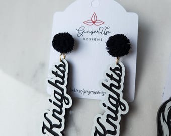 May include: Black pom-pom earrings with gold-coloured hooks. The earrings feature a white base with the word "Knights" in a black, cursive font. The earrings are displayed on a white card with the "Ginger Up Designs" logo.