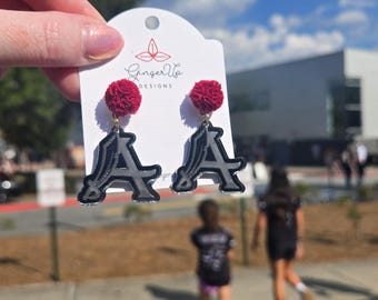 May include: Earrings with a dark grey letter "A" design and a black outline, paired with a small, round, red pom-pom. The earrings are presented on a white card with the text "Ginger Up Designs".