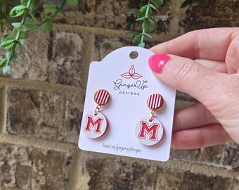 May include: Red and white letter "M" earrings. The earrings have a circular design with a red and white striped top and a white circle with a red "M" in the centre. The earrings are held up by a person.