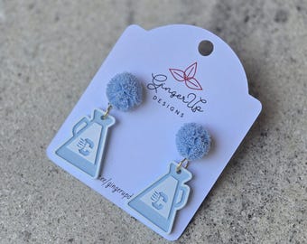 May include: Pale blue earrings featuring a megaphone design and a fluffy blue pom-pom. The earrings are attached to a white card with the text "Ginger Up Designs" and a floral logo. The earrings are on a grey surface.