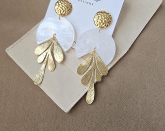 May include: Gold-coloured earrings with a textured disc top, a white semi-circle, and a leaf-shaped drop. The earrings are displayed on a cream-coloured card with the text "Ginger Up Designs". The background is a neutral tan.
