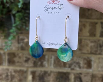 May include: Teardrop earrings with gold-coloured hooks, displaying a marbled pattern in blue and green. The earrings are presented on a white tag with the text "Ginger Up Designs" and a website link. The earrings are a stylish accessory.