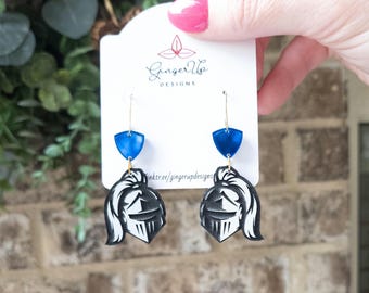 May include: Dangle earrings with a blue shield and a black and white knight's helmet design. The earrings hang from gold-coloured hooks. The card behind the earrings has the text "Ginger Up Designs".