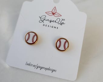 May include: Baseball stud earrings. Each earring has a white baseball design with red stitching, set in a gold-coloured metal frame. The earrings are displayed on a white card with the text "Ginger Up Designs" and a website link.