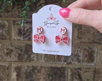 May include: Red bow earrings with a white border and a gold-coloured circle with the letter "M" inside. The earrings are displayed on a white card with the text "Ginger Up Designs" and a link to their website.