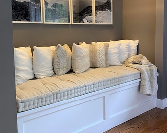 May include: A white window seat with a beige cushion and pillows. The seat is built into a wall and has a white trim. There are four framed black and white photos above the seat.