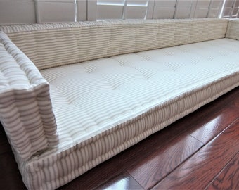 May include: A long, rectangular, white and gray striped cushion with a tufted top. The cushion is made of fabric and has a thick, padded base. It is sitting on a wooden floor.