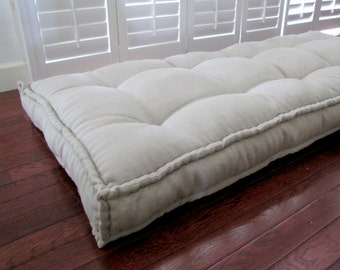 May include: A large, off-white floor cushion with a tufted design and a thick, stitched border. The cushion is made of a soft, linen-like fabric and is perfect for adding comfort and style to any room.