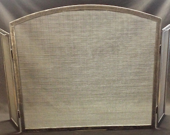 May include: A silver metal fireplace screen with a mesh center panel and two hinged side panels. The screen has an arched top and a rectangular base. The mesh is a fine weave, and the frame is a brushed metal finish.