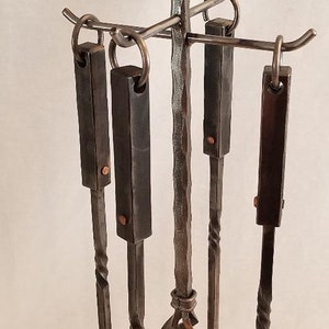 Pu&ograve; includere: Un set di cinque strumenti per camino in metallo nero con un design attorcigliato. Gli strumenti sono appesi a un supporto con una parte superiore rotonda. Gli strumenti includono un attizzatoio, delle pinze, una pala, una spazzola e un supporto.