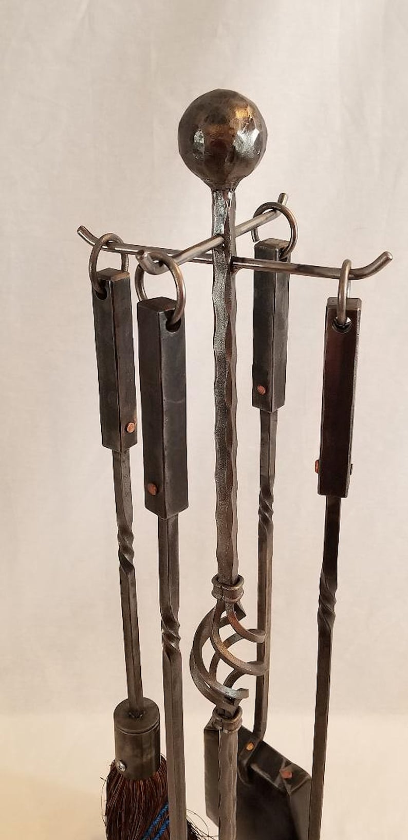 Pu&ograve; includere: Un set di cinque strumenti per camino in metallo nero con un design attorcigliato. Gli strumenti sono appesi a un supporto con una parte superiore rotonda. Gli strumenti includono un attizzatoio, delle pinze, una pala, una spazzola e un supporto.
