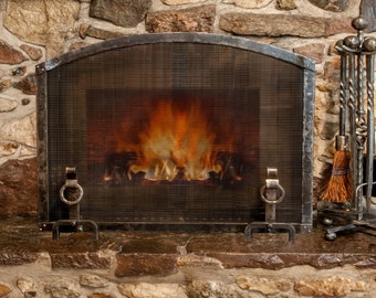 May include: A black wrought iron fireplace screen with an arched top and a fine mesh center. The screen features two decorative ring handles at the base and stands in front of a burning fire. A matching set of fireplace tools is positioned to the right.