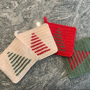 Op de afbeelding: Vier gehaakte kerstboom pannenlappen in crème, rood en groen. Elke vierkante pannenlap heeft een boomontwerp in een contrasterende kleur. De pannenlappen hebben een lus om op te hangen en liggen op een gespikkeld grijs oppervlak.