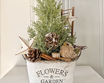 May include: A decorative arrangement in a white bucket with the text "FLOWERS & GARDEN". It features a small evergreen tree, star ornaments, a pine cone, and a bird figurine. The bucket is on a white surface.