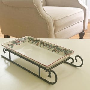 May include: A rectangular ceramic serving tray with a cream-colored center and a border featuring green pine branches, red berries, and colorful ornaments. The tray rests on a dark metal stand shaped like a sled with curved runners.