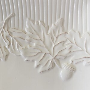 May include: White ceramic serving dish with a raised design of oak leaves and acorns. The dish has a textured surface with vertical lines.