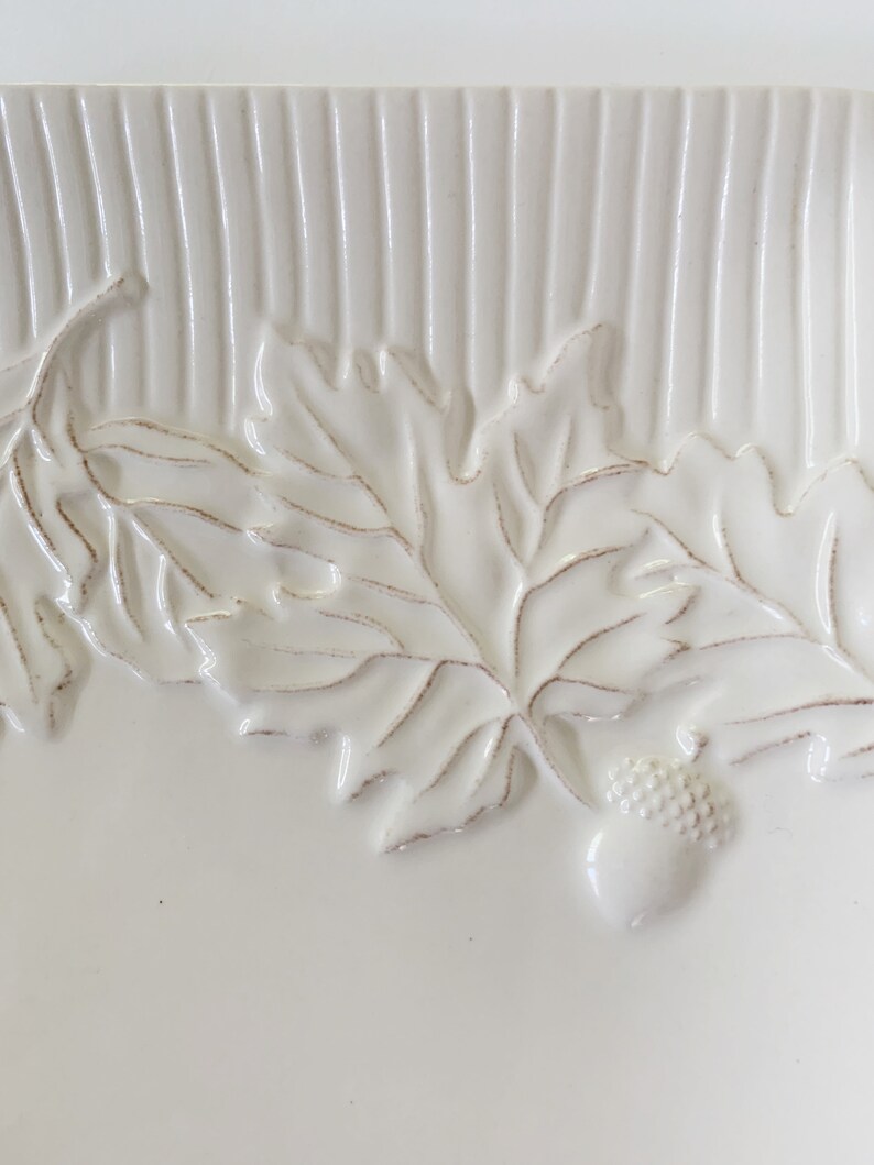 May include: White ceramic serving dish with a raised design of oak leaves and acorns. The dish has a textured surface with vertical lines.