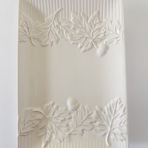 May include: White ceramic serving dish with an embossed leaf and acorn design. The dish has a rectangular shape and a slightly raised rim.