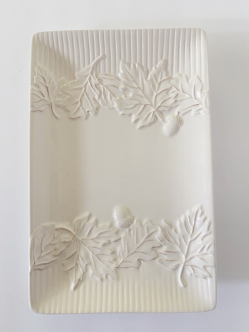 May include: White ceramic serving dish with an embossed leaf and acorn design. The dish has a rectangular shape and a slightly raised rim.