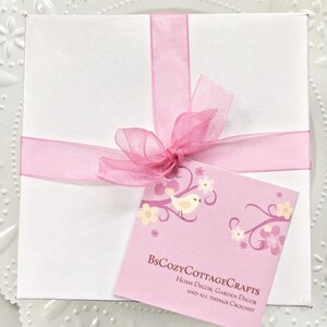 May include: A white gift box tied with a pink ribbon and bow. A pink tag with a bird and floral design is attached to the box. The tag reads "BsCozyCottageCRAFTS Home Decor, Garden Decor and all things Crochet".