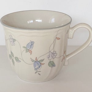 Puede incluir: Una taza de cerámica blanca con un diseño floral con flores azules y rosas y hojas verdes. La taza tiene una forma redondeada y un asa.