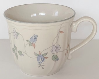 May include: A white ceramic mug with a floral design featuring blue and pink flowers and green leaves. The mug has a rounded shape and a handle.