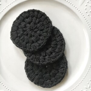 May include: Three black crocheted makeup remover pads on a white plate.
