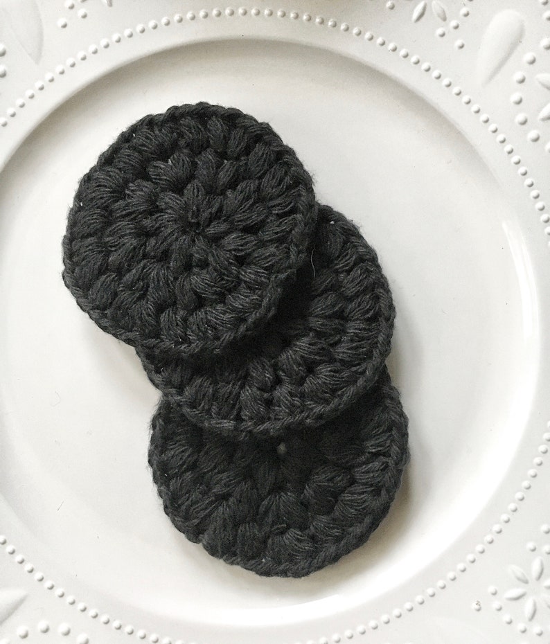 May include: Three black crocheted makeup remover pads on a white plate.