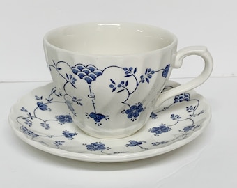 May include: A white ceramic teacup and saucer set featuring a delicate blue floral pattern with small blossoms, vines, and scalloped edges. The teacup has a ribbed texture and a curved handle, while the saucer matches the floral design.