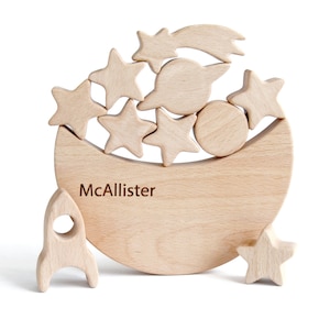 Wooden moon stacking and balance game in natural beech wood, crescent moon rocker holding six stars, a shooting star comet, planet and rocket, unpainted Waldorf style fine motor toy for toddlers, space theme nursery shelf decor, white background.